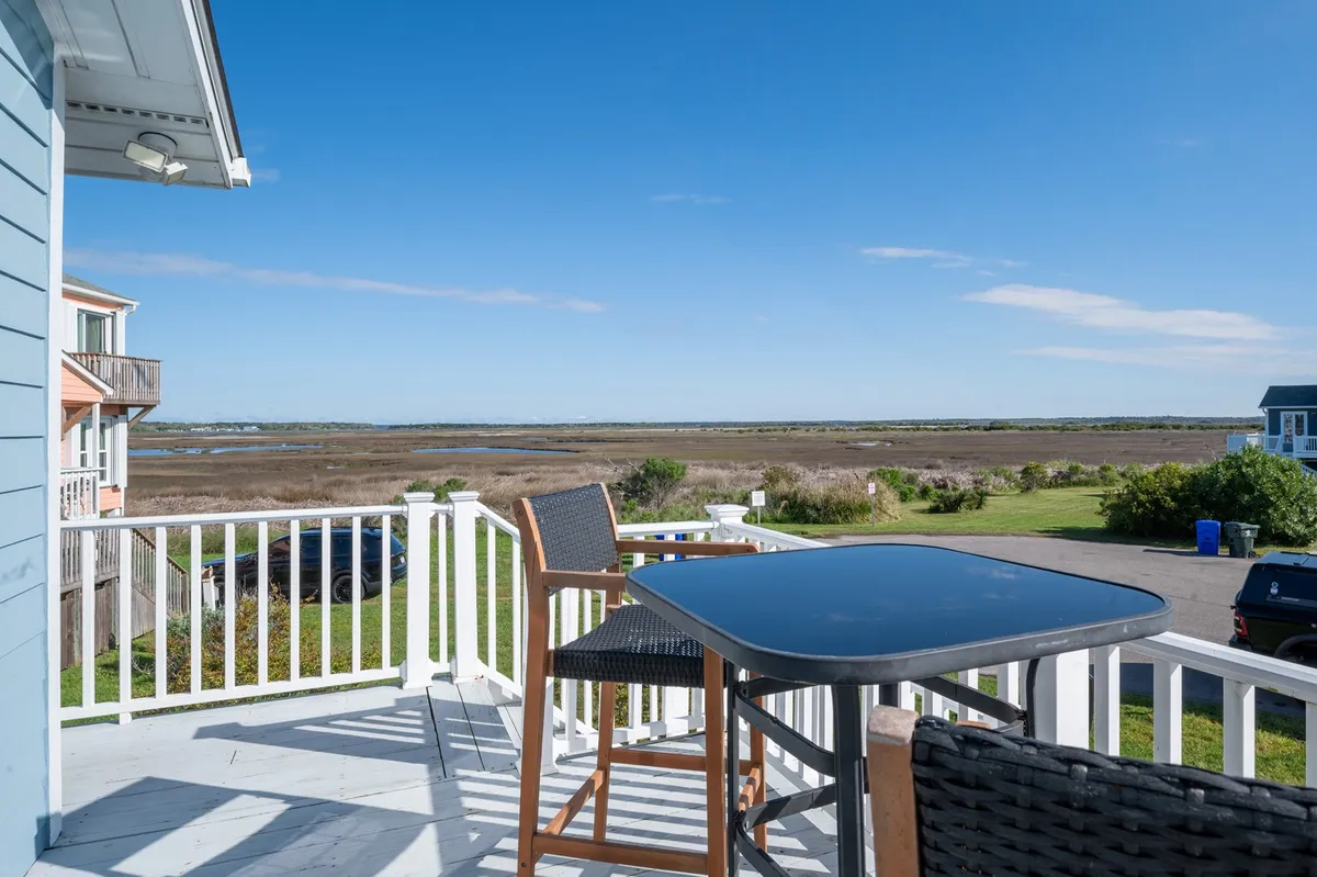Deck furniture is arranged for relaxation, facing a wide view of the marsh under a blue sky. The space invites guests to relax with a drink, observe wildlife, or enjoy stunning sunsets, capturing the peaceful and immersive setting of the back porch.