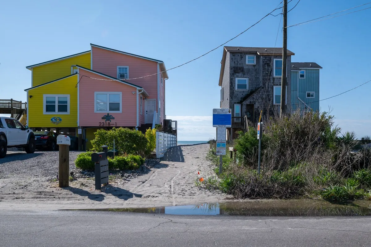 A cheerful scene featuring a pathway flanked by colorful coastal houses leading directly to the beach, with vibrant local vegetation and clear signage in view. The image evokes the atmosphere of a welcoming, walkable neighborhood on the coast.