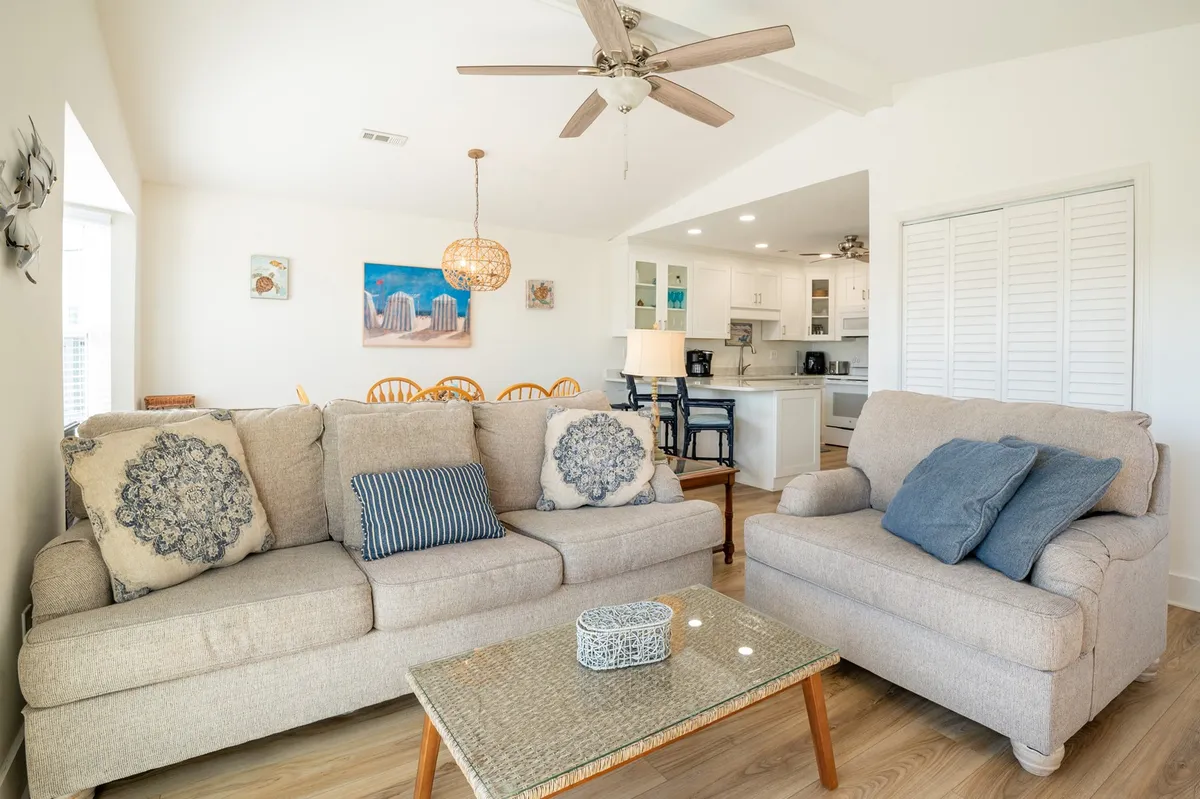 An inviting open-concept living space with plush sofas adorned with decorative cushions, a glass coffee table, and a dining area visible in the background. The space is bathed in natural light, offering a comfortable spot for relaxing, socializing, or entertaining guests.