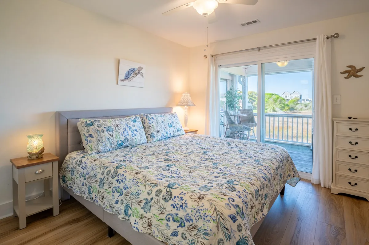This image features a queen bed with floral bedding, a sliding glass door that opens to the outdoors, and a nightstand with a lamp. The room feels light, airy, and inviting, with a focus on comfort and relaxation.