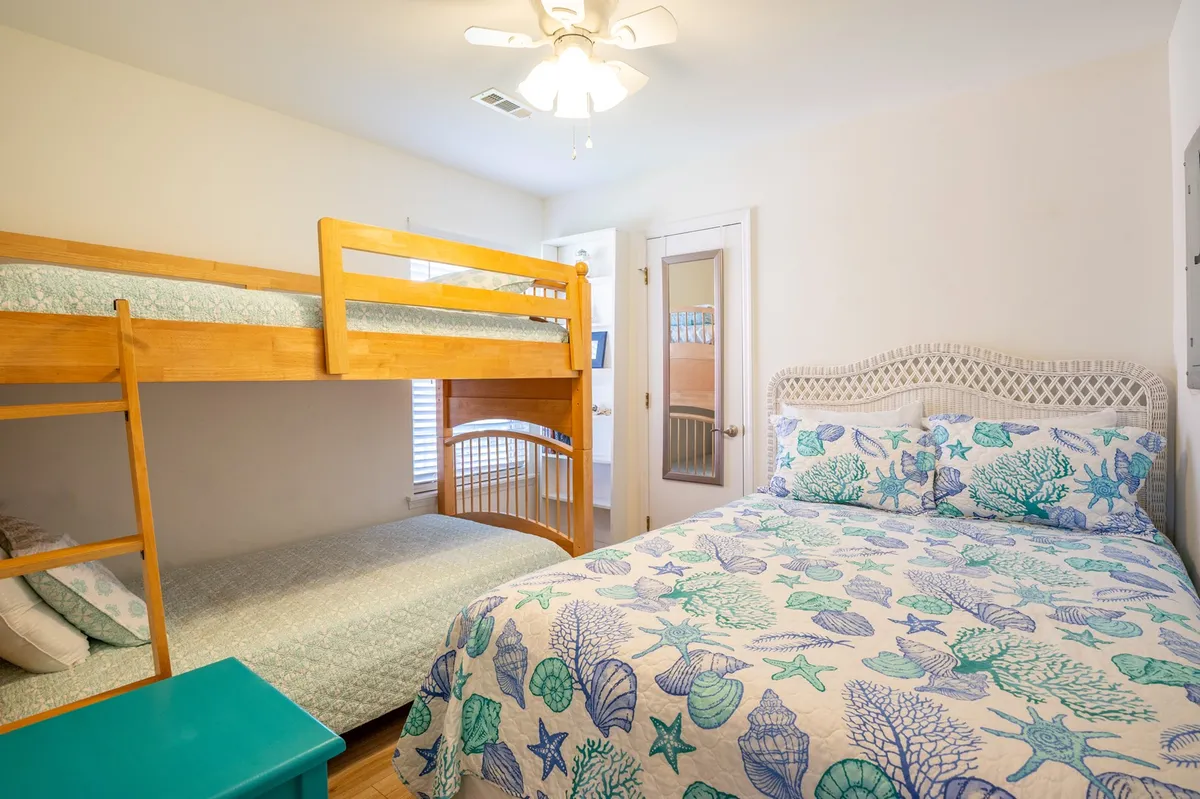 The room contains a cheerful bunk bed and a queen bed with a decorative quilt, creating a playful and cozy environment. Sunlight pours through the windows, setting a relaxed and inviting mood ideal for families or children.