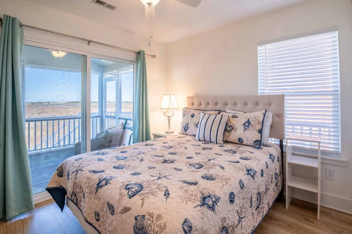 The image displays a serene bedroom with seashell-patterned bedding, large windows, and doors that open directly onto a balcony. A bedside table with a lamp is positioned next to the bed, and natural light fills the space, suggesting a calming retreat with immediate outdoor access.