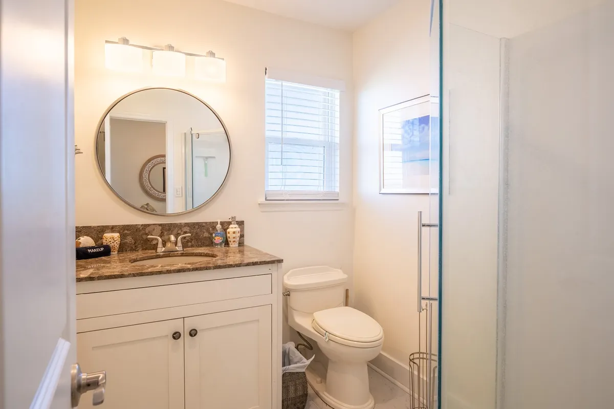 A modern bathroom featuring a circular mirror, granite countertop, and a glass shower. The space is bright, tranquil, and designed for relaxation and self-care.