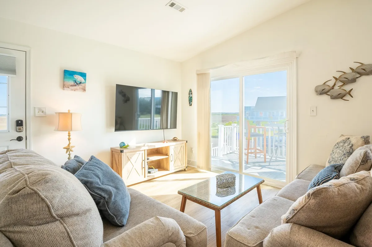 A cozy living room with a sofa and cushions facing a television, complemented by a coffee table and sliding glass doors leading to a balcony. The abundance of natural light and easy outdoor access create a bright, airy environment ideal for relaxation and family activities.
