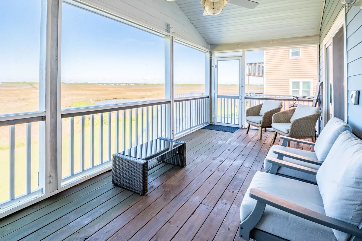 This image features a comfortable seating area with a glass table on a screened porch, offering a spacious view beyond. The space is set up for relaxing with a book, enjoying morning coffee, or socializing with friends. The casual and serene mood is complemented by natural light and sightlines to the outdoors.