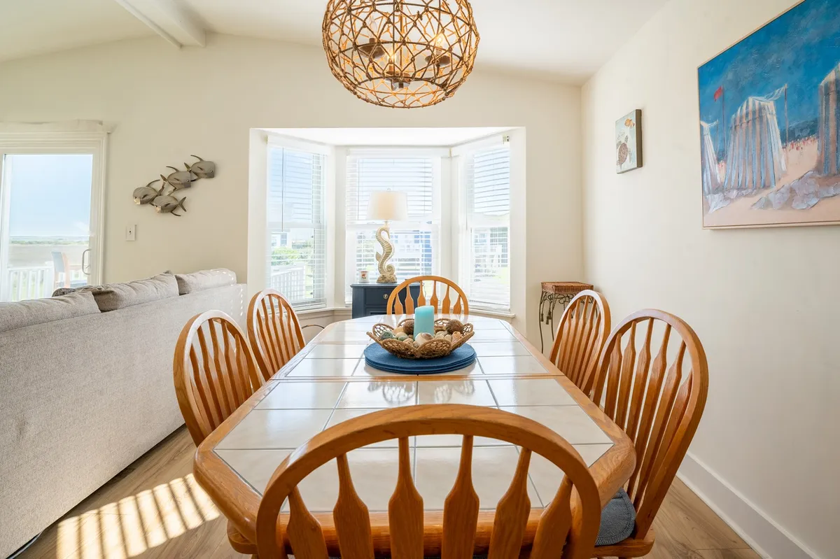 A warmly lit dining area with a welcoming dining table under a chandelier, a bay window allowing natural light, and a decorative painting on the wall. The table is set for gatherings, hinting at family meals, social events, and game nights.