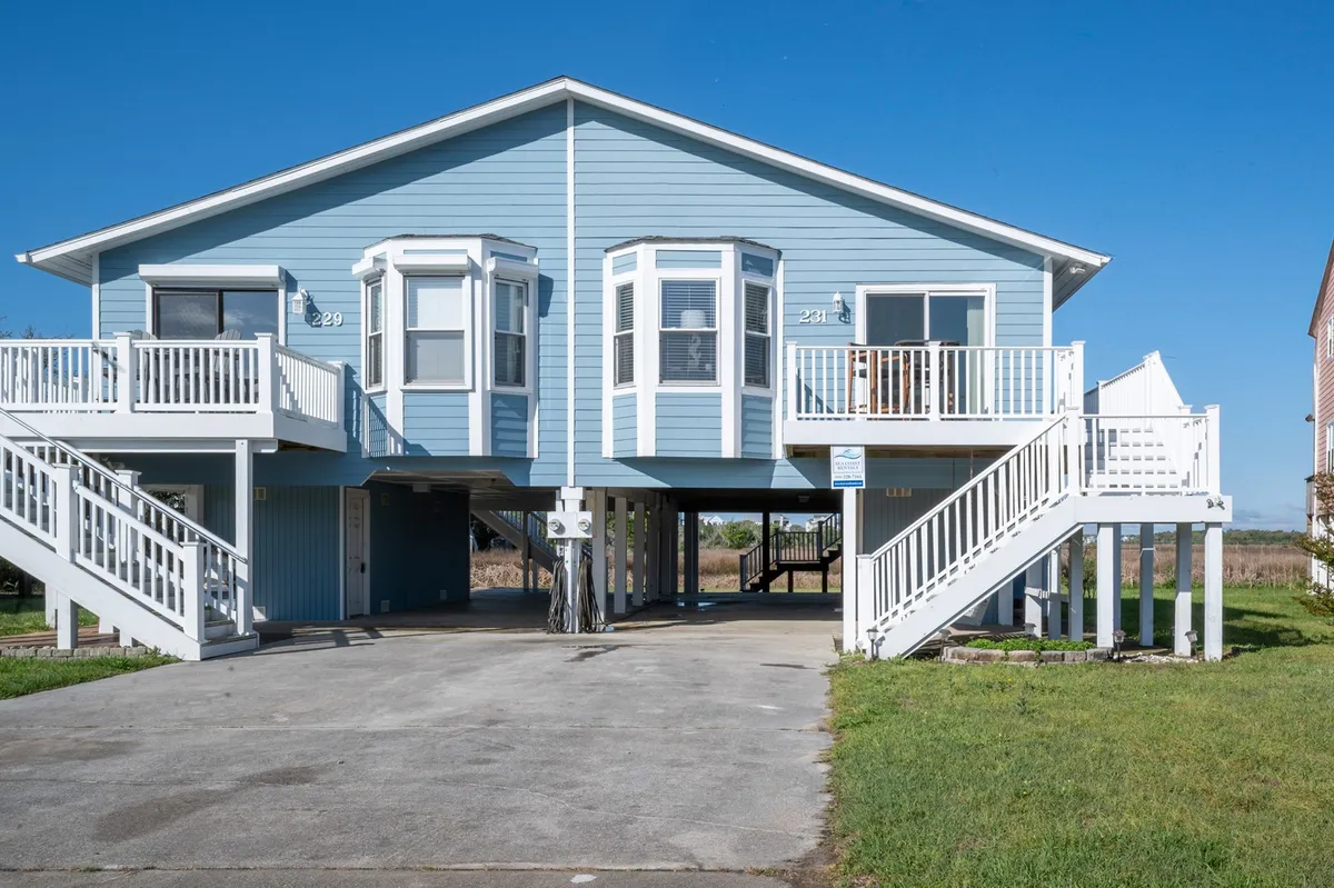 A broad exterior view showcasing the blue house with bay windows, dual staircases leading to entrances, and space suitable for family gatherings and outdoor barbecues, all in a lush, marsh-adjacent setting.