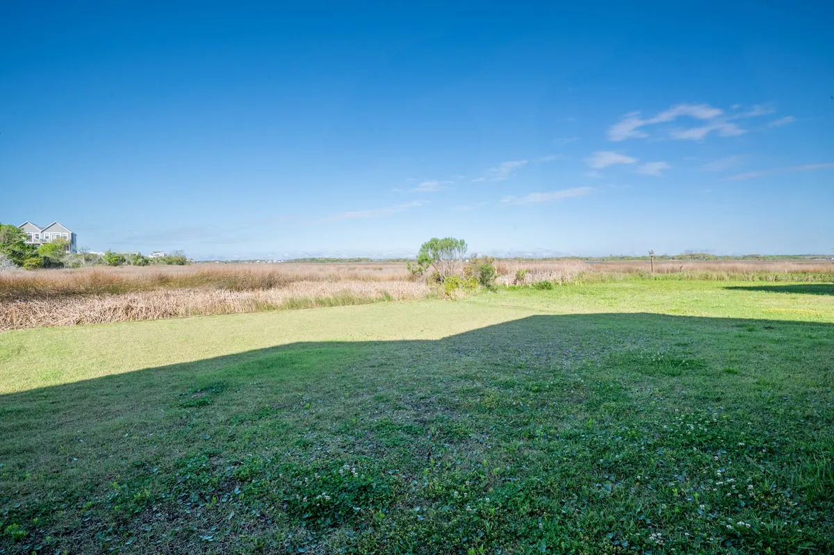 The image showcases an open green space leading to distant marshland, with a clear blue sky overhead. It sets a peaceful, natural backdrop ideal for birdwatching, sunrise appreciation, and immersion in the marsh landscape.